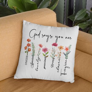 Pode incluir: Travesseiro branco com um design floral e o texto "God says you are" com as palavras "&uacute;nico, especial, ador&aacute;vel, precioso, forte, escolhido, perdoado" escritas verticalmente abaixo do texto.