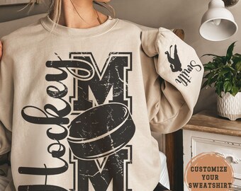 May include: Beige sweatshirt with the words "Hockey Mom" in black, featuring a hockey puck graphic. The sleeve has a hockey player silhouette and the name "Smith". The sweatshirt has a distressed look. The text "Customize Your Sweatshirt" is also visible.