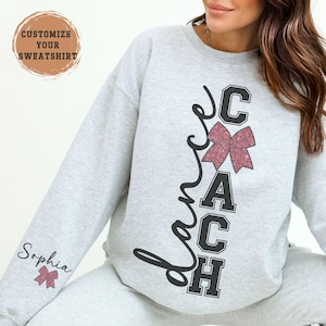 May include: A light gray sweatshirt with the words "Dance Coach" in black script and block letters. A pink glitter bow is in the center. The sleeve has the name "Sophia" and a pink bow. The top left corner has the text "Customize Your Sweatshirt".