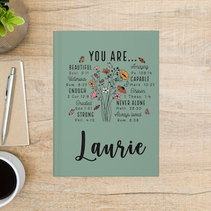 May include: A sage green journal with the words "YOU ARE..." and inspirational affirmations, including "Beautiful," "Victorious," and "Strong." A floral bouquet and the name "Laurie" are also featured. A pen and coffee cup are in the background.