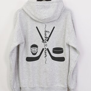 K&ouml;nnte beinhalten: Grauer Kapuzenpullover mit einem Hockey-Schl&auml;ger- und Puck-Design. Der Text "hockey & mom" ist vertikal auf der R&uuml;ckseite des Pullovers geschrieben.