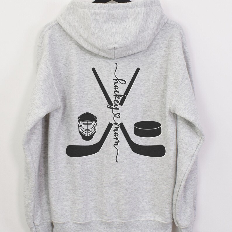 K&ouml;nnte beinhalten: Grauer Kapuzenpullover mit einem Hockey-Schl&auml;ger- und Puck-Design. Der Text "hockey & mom" ist vertikal auf der R&uuml;ckseite des Pullovers geschrieben.