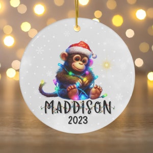 May include: A white ceramic Christmas ornament with a cartoon monkey wearing a Santa hat and holding colorful Christmas lights. The ornament is personalized with the name "MADDISON" and the year "2023".