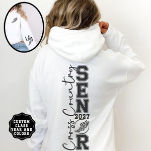 May include: A white hooded sweatshirt featuring black text on the back that reads Cross Country SENIOR 2027 with a winged graphic. The sleeve displays the name Lily and a small winged icon. The design includes custom class year and colors.