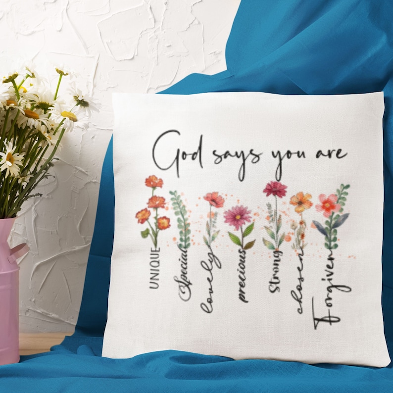 Pode incluir: Travesseiro branco com o texto "Deus diz que voc&ecirc; &eacute;" e um desenho floral com as palavras "&uacute;nico, especial, ador&aacute;vel, precioso, forte, escolhido, perdoado" escritas verticalmente abaixo das flores.