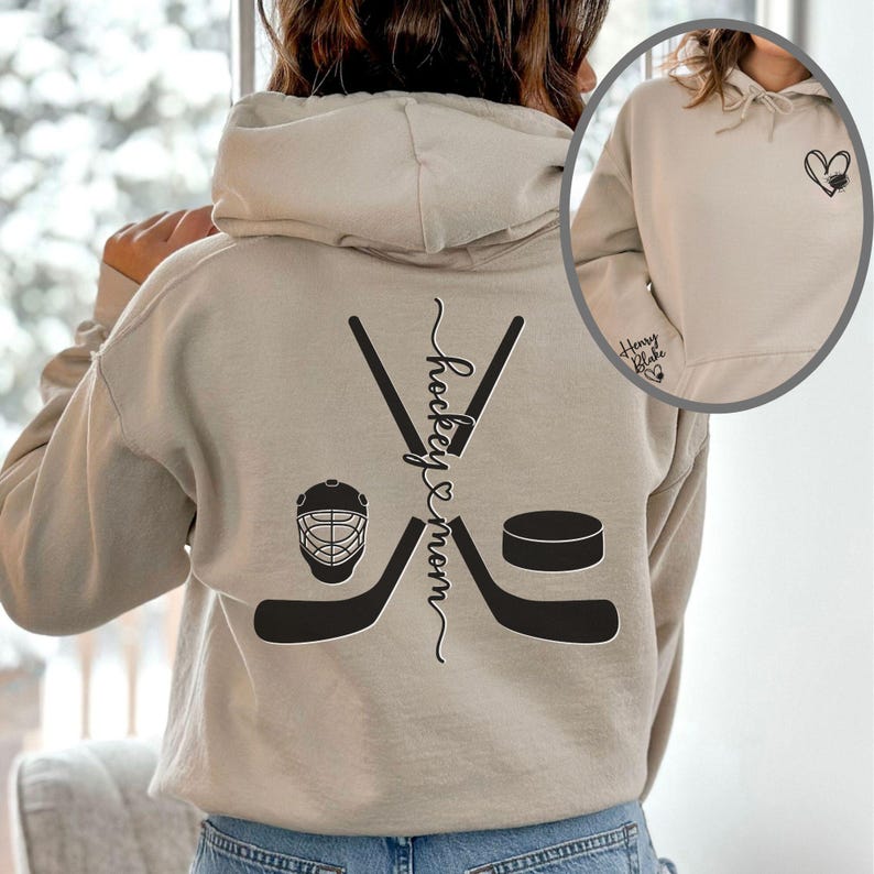 K&ouml;nnte beinhalten: Ein beiger Hoodie mit einem schwarzen Hockey-Schl&auml;ger- und Puck-Design auf der R&uuml;ckseite. Der Text "hockey mom" ist vertikal auf der R&uuml;ckseite des Hoodies geschrieben. Die Vorderseite des Hoodies hat ein kleines schwarzes Herz mit dem Text "Hockey Babe" auf der linken Seite.
