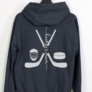 K&ouml;nnte beinhalten: Ein dunkelblauer Kapuzenpullover mit einer wei&szlig;en Grafik von gekreuzten Hockeyschl&auml;gern, einem Puck und einem Hockeyhelm. Der Text "hockey & mom" ist vertikal zwischen den Schl&auml;gern geschrieben.