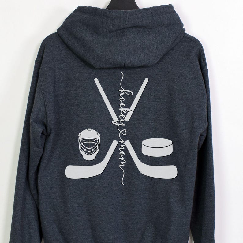 K&ouml;nnte beinhalten: Ein dunkelblauer Kapuzenpullover mit einer wei&szlig;en Grafik von gekreuzten Hockeyschl&auml;gern, einem Puck und einem Hockeyhelm. Der Text "hockey & mom" ist vertikal zwischen den Schl&auml;gern geschrieben.