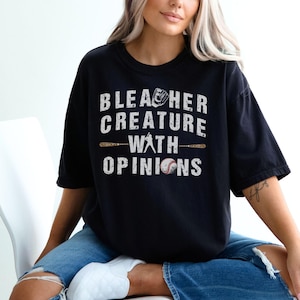 May include: Black t-shirt with the text "Bleacher Creature With Opinions" in white. The design includes a baseball glove, baseball bats, and a baseball. The shirt is a casual, oversized fit.