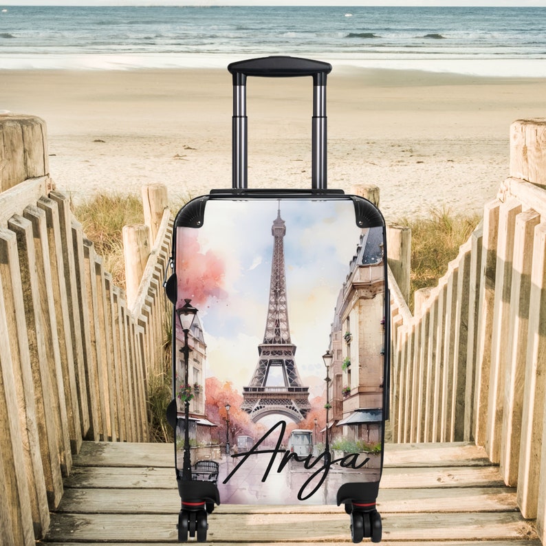 May include: A black suitcase with a watercolor design of the Eiffel Tower in Paris, France. The suitcase has a retractable handle and wheels. The name "Anya" is written on the suitcase.