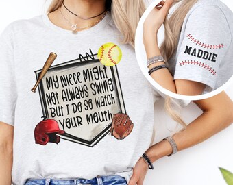 May include: A gray t-shirt with a baseball-themed design. The design features a baseball diamond, a baseball bat, a baseball glove, a baseball helmet, and a softball. The text on the shirt reads "An my niece might not always swing but I do so watch your mouth". The shirt also has a personalized name on the sleeve.