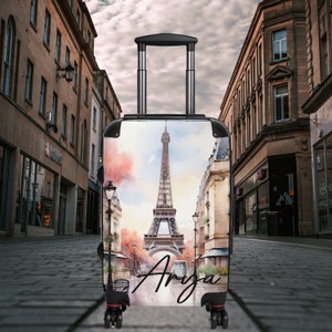 May include: A black carry-on suitcase with a watercolor design of the Eiffel Tower in Paris, France. The suitcase has a black handle and wheels. The name "Anya" is written on the front of the suitcase.