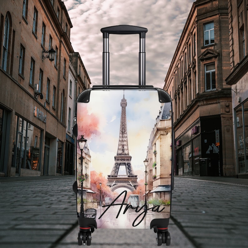 May include: A black carry-on suitcase with a watercolor design of the Eiffel Tower in Paris, France. The suitcase has a black handle and wheels. The name "Anya" is written on the front of the suitcase.