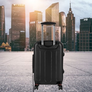 May include: A black suitcase with a retractable handle and four wheels. The suitcase is standing on a gray concrete surface in front of a city skyline.