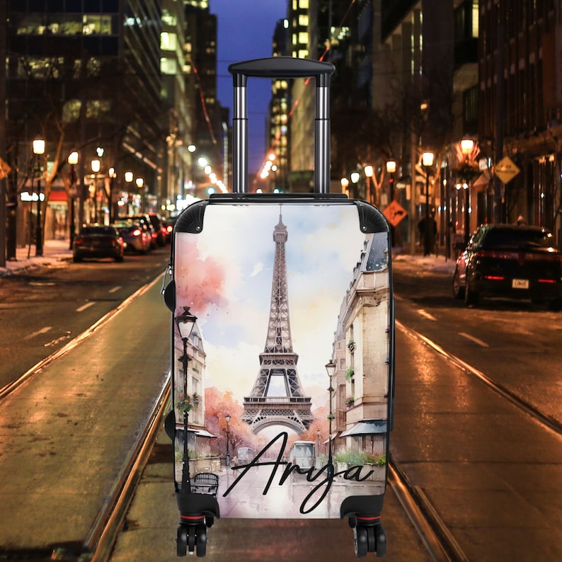 May include: A white suitcase with a watercolor design of the Eiffel Tower in Paris, France. The suitcase has a black handle and wheels. The name "Anya" is written in cursive on the front of the suitcase.