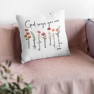 Pode incluir: Travesseiro branco com o texto "God says you are" e uma lista vertical de palavras: "&uacute;nico, especial, ador&aacute;vel, precioso, forte, escolhido, perdoado". As palavras s&atilde;o cercadas por flores e vegeta&ccedil;&atilde;o em aquarela.