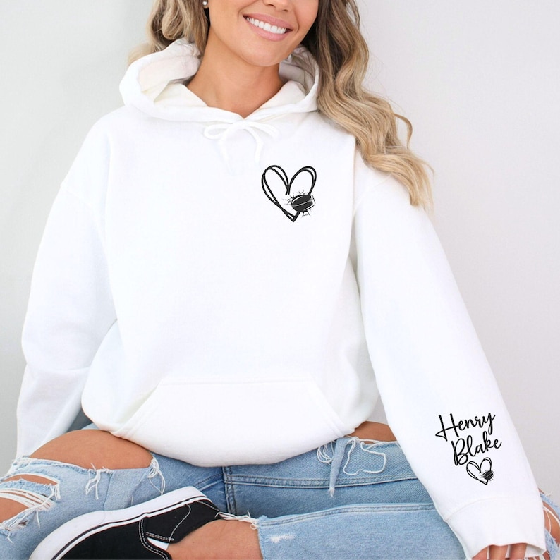K&ouml;nnte beinhalten: Wei&szlig;er Hoodie mit einem schwarzen Herzdesign auf der Vorderseite und dem Text "Henry Blake" mit einem Herz auf dem &Auml;rmel.