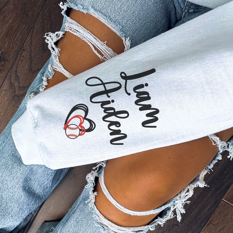 May include: A light blue sweatshirt with the text "Liam Hidden" in black cursive lettering and a red and white baseball with two red hearts above it.