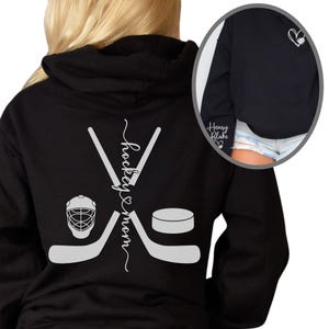 K&ouml;nnte beinhalten: Schwarzer Hoodie mit einer wei&szlig;en Grafik von Hockeyschl&auml;gern, einem Puck und einem Helm. Der Text "hockey mom" ist vertikal auf der R&uuml;ckseite des Hoodies geschrieben.