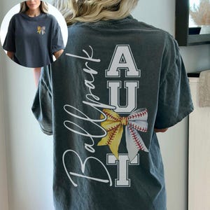 May include: Dark grey t-shirt with a baseball-themed design. The back features the text "Ballpark" in a script font, with "AUT" in block letters and a baseball bow graphic. A smaller version of the bow is on the front.
