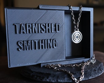 May include: A silver necklace with a circular pendant featuring concentric circles hangs inside a black box. The box is labeled "Tarnished Smithing" in raised letters. The box sits on a dark, textured surface.