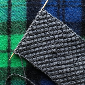 May include: A close-up of a grey knitted fabric with a textured stitch pattern. The fabric is being worked on with a wooden knitting needle and grey yarn.