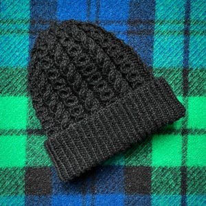 May include: A dark grey knitted beanie hat with a textured pattern and a folded brim. The hat is made of a soft, warm material, ideal for cold weather. The hat is displayed on a plaid background.