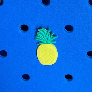 May include: A yellow pineapple charm with green leaves on a blue background with a pattern of holes.