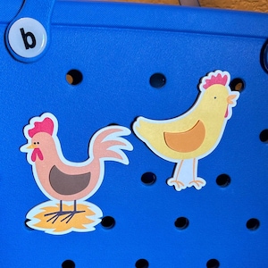 May include: Two colorful chicken stickers on a blue background. The rooster is brown with a red comb and wattle. The hen is yellow with a red comb and wattle. Both stickers have a white outline.