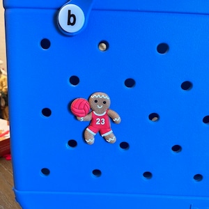 May include: A blue tote bag with a gingerbread man charm wearing a red jersey with the number 23 and holding a basketball. The charm is attached to the bag with a white button that says "b".
