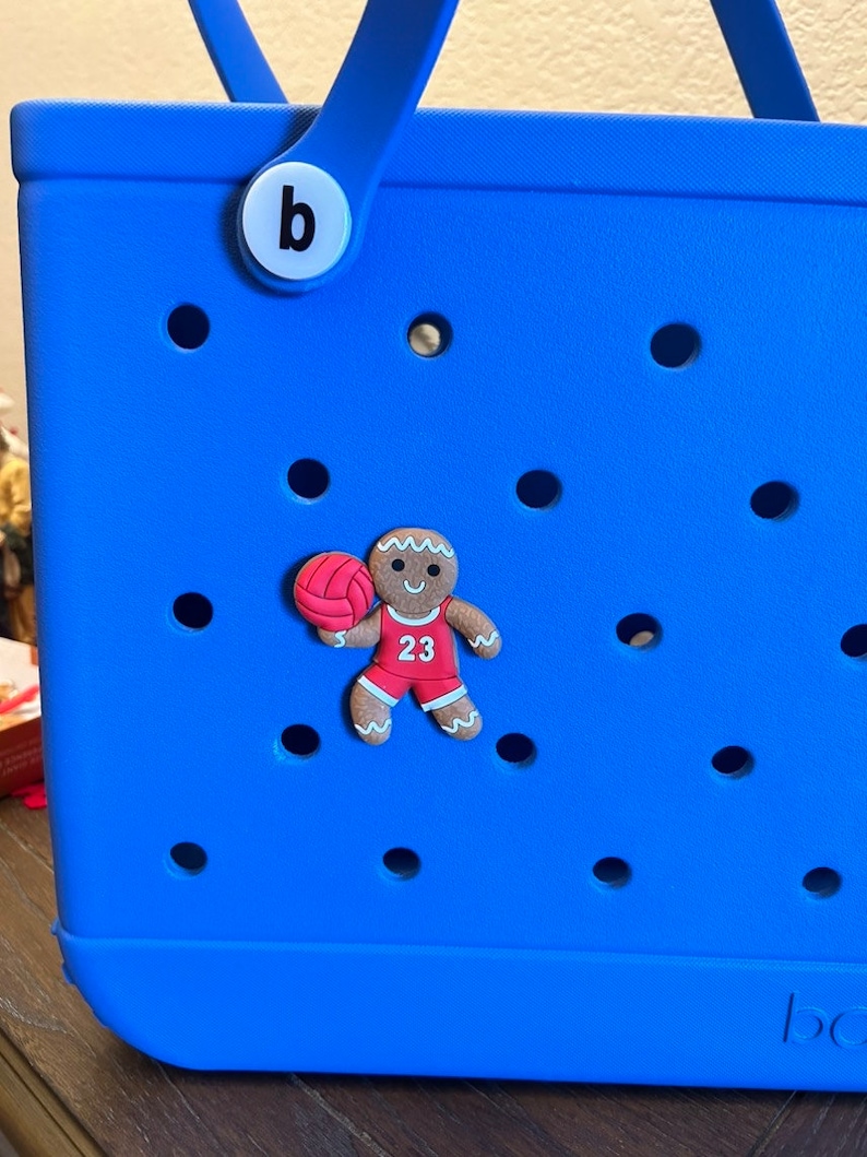 May include: A blue tote bag with a gingerbread man charm wearing a red jersey with the number 23 and holding a basketball. The charm is attached to the bag with a white button that says "b".