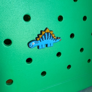 May include: A blue dinosaur charm with yellow accents is affixed to a bright green surface with several round holes. The dinosaur has a friendly expression and is likely a decorative accessory.