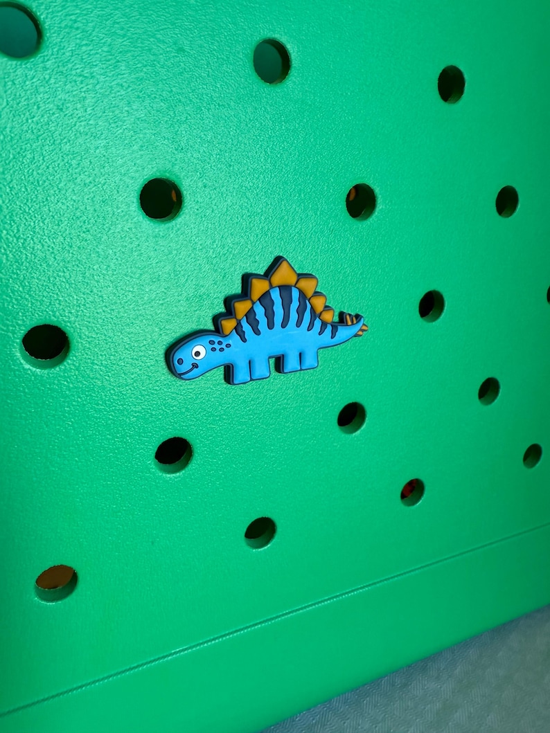May include: A blue dinosaur charm with yellow accents is affixed to a bright green surface with several round holes. The dinosaur has a friendly expression and is likely a decorative accessory.