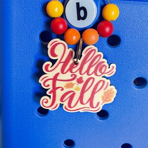 May include: A blue and white charm with the text "Hello Fall" in red cursive lettering. The charm has a small metal ring and is attached to a string of colorful beads.