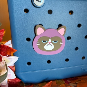 May include: A blue tote bag with a grumpy cat charm attached. The charm features a brown cat face with a pink hood. The bag has a handle and a series of circular holes. The background includes fall leaves.