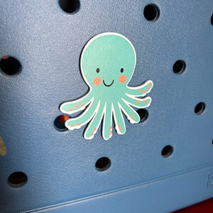May include: A blue tote bag with a smiling octopus sticker. The octopus is light green with pink cheeks and orange spots.