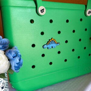 May include: A bright green tote bag with a dinosaur-shaped charm. The bag has a series of circular cutouts and two handles. The dinosaur charm is blue with yellow and orange accents. The bag is made of a durable material.