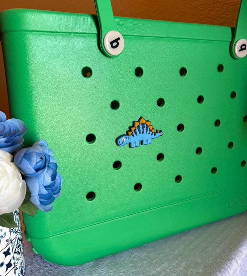 May include: A bright green tote bag with a dinosaur-shaped charm. The bag has a series of circular cutouts and two handles. The dinosaur charm is blue with yellow and orange accents. The bag is made of a durable material.