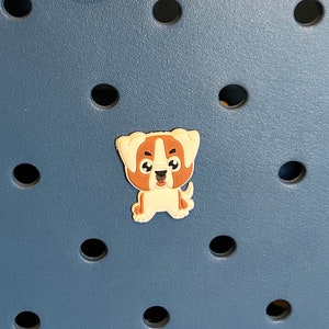 Op de afbeelding: Een kleine, cartoonachtige hondenbedel in bruin en wit. De bedel staat tegen een blauwe achtergrond met gelijkmatig verdeelde zwarte cirkelvormige gaten. De hond heeft een vriendelijke uitdrukking.