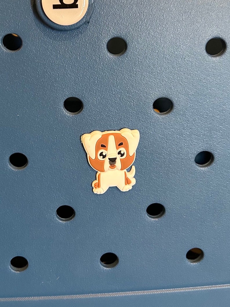 Op de afbeelding: Een kleine, cartoonachtige hondenbedel in bruin en wit. De bedel staat tegen een blauwe achtergrond met gelijkmatig verdeelde zwarte cirkelvormige gaten. De hond heeft een vriendelijke uitdrukking.
