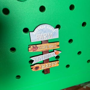May include: A green wooden sign with the words "Harvest Acres Farm" on it. The sign is attached to a wooden post with three smaller signs attached to it. The smaller signs say "Corn Maze", "Hayrides", and "Happy".