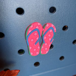 May include: A pair of pink flip-flops with white polka dots, blue straps, and green edges. The flip-flops are placed on a blue surface with several black circular holes.