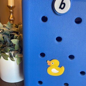 K&ouml;nnte beinhalten: Eine blaue Tragetasche mit einem gelben Gummiente-Anh&auml;nger und einem wei&szlig;en Knopf mit dem Buchstaben "b" darauf.