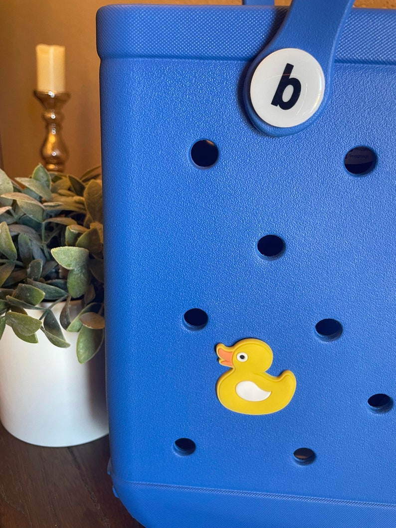 K&ouml;nnte beinhalten: Eine blaue Tragetasche mit einem gelben Gummiente-Anh&auml;nger und einem wei&szlig;en Knopf mit dem Buchstaben "b" darauf.