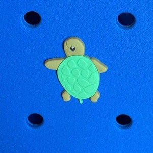 May include: A green and brown turtle charm with a textured shell. The turtle is on a blue background.