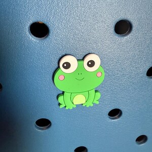 May include: A green frog with white eyes and pink cheeks is attached to a blue surface with holes.