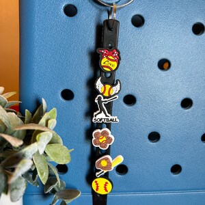 May include: A black charm bracelet with a variety of softball-themed charms, including a softball with wings, a glove and bat, a flower with a softball in the center, and a softball with the word "Love" on it.