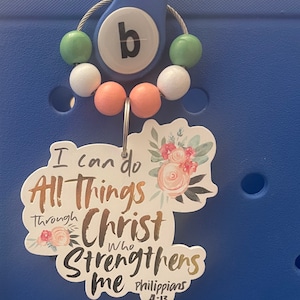 May include: A white and gold charm with the text "I can do all things through Christ who strengthens me Philippians 4:13" with floral accents. The charm is attached to a blue tote bag with a silver ring and colorful beads.