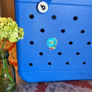 May include: A blue tote bag with a white letter "b" on the handle and a round sticker that says "Best Teacher Ever" with a rainbow design. The bag has a pattern of small holes.
