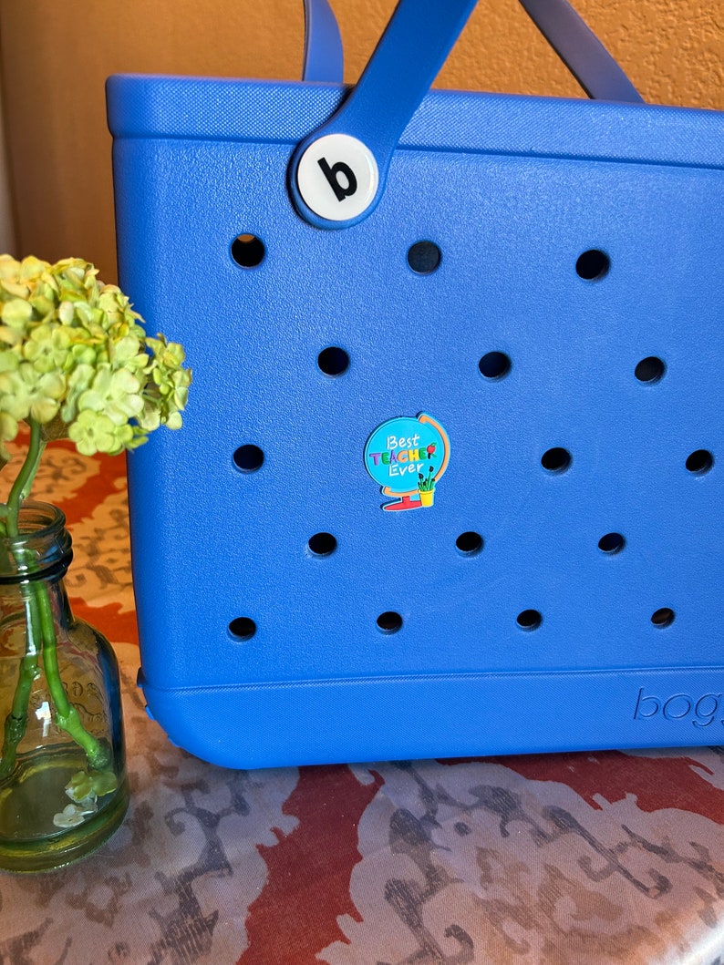 May include: A blue tote bag with a white letter "b" on the handle and a round sticker that says "Best Teacher Ever" with a rainbow design. The bag has a pattern of small holes.